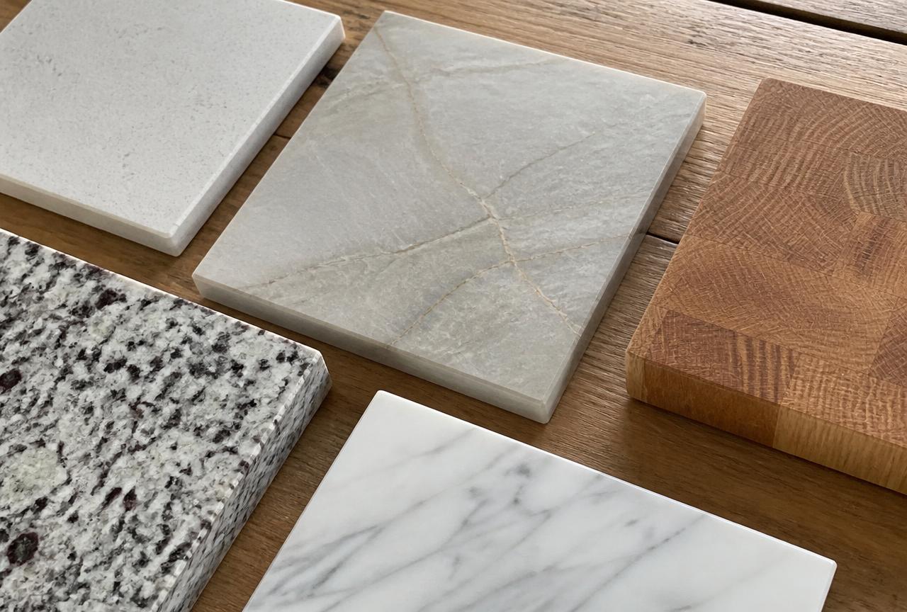 Kitchen countertop samples including quartz, quartzite, granite, marble, and butcher block on a wood surface